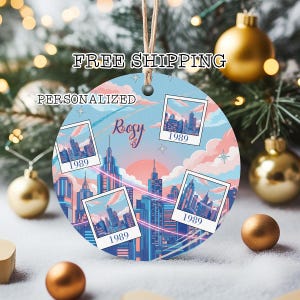 May include: A round Christmas ornament with a cityscape design in shades of blue, pink, and orange. The ornament features four framed images of the city and the year 1989. The name "Rosy" is written in cursive. A string is attached for hanging.