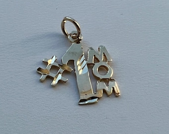 May include: A gold-colored charm pendant with the words "#1 MOM". The charm features a hashtag symbol, the number one, and the word "MOM" in capital letters. The pendant has a small loop at the top for attaching to a chain or bracelet.