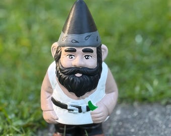 May include: A garden gnome with a black hat, a white shirt, and a black beard. The gnome is holding a green leaf in its hand.