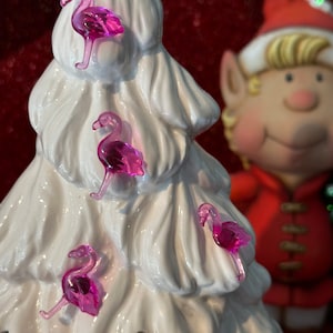 May include: A white ceramic Christmas tree decorated with pink flamingo ornaments. The tree is in front of a figurine of an elf wearing a red and white Santa hat.