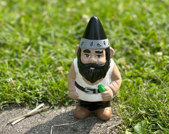 May include: A black and white garden gnome with a black pointed hat and a grey band around the hat. The gnome has a black beard and is holding a green object in its right hand.