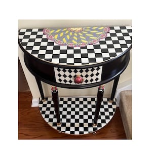 May include: A black and white checkered half-moon console table with a drawer. The table features a decorative top with a geometric design and a red and gold knob. The legs are black with gold accents.