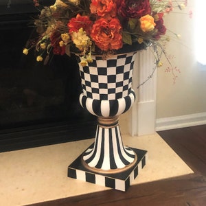 May include: A black and white checkered ceramic vase with a gold trim and a black and white striped base. The vase is sitting on a white pedestal and is filled with a bouquet of orange, yellow, and red flowers.