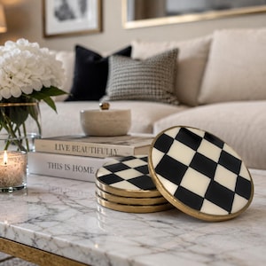 May include: A stack of four decorative coasters with a black and white checkered pattern and gold-colored edges. The coasters are on a marble coffee table, with a vase of white flowers, books, and a lit candle in the background.