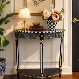 May include: A decorative half-moon console table with black and white striped legs and a mosaic tile border. The table features a black top and a lower shelf. A lamp, books, and a flower arrangement sit on top.