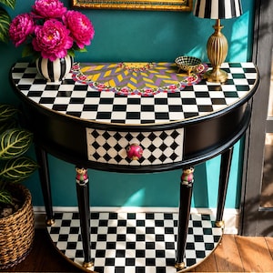 May include: A black and white checkered console table with a drawer and a decorative top. The table has a pink knob, gold accents, and a lower shelf. A vase of pink flowers and a striped lamp sit on top.