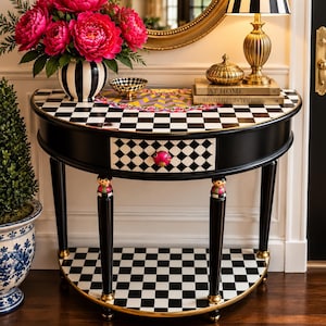 May include: A semi-circular console table featuring a black and white checkered pattern on the top and bottom shelves. The table has a single drawer with a pink knob, black legs with decorative figures, and gold-toned accents.