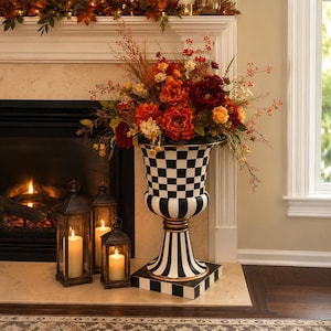 May include: A decorative urn with a black and white checkered pattern, filled with an arrangement of fall-colored flowers. Three black lanterns with lit candles sit in front of a fireplace, creating a warm, inviting ambiance.