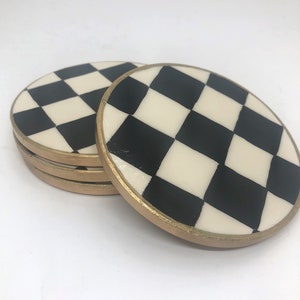 Può includere: Set di quattro sottobicchieri rotondi con un motivo a scacchiera bianco e nero. I sottobicchieri hanno un bordo dorato.