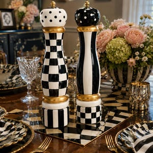 May include: Two decorative salt and pepper grinders with gold accents. One is black and white checkered, the other is black and white striped. Both have gold bands and dotted or striped tops. The grinders are on a table set for a formal dinner.