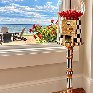 May include: A tall gumball machine featuring a clear glass globe filled with red candies. The base and stand are decorated with a black and white checkered pattern, accented by ornate gold metal details and a gold coin mechanism.