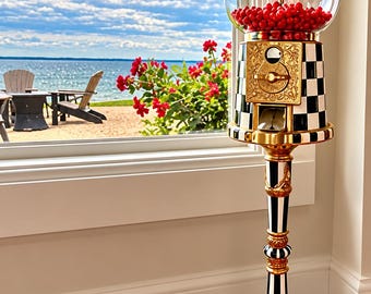 May include: A tall gumball machine featuring a clear glass globe filled with red candies. The base and stand are decorated with a black and white checkered pattern, accented by ornate gold metal details and a gold coin mechanism.