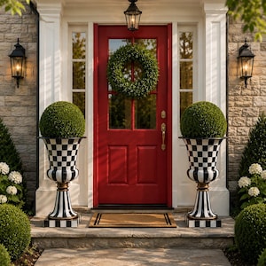 May include: A red front door with a green wreath, flanked by two black and white checkered urns holding green topiary balls. The entrance is framed by white columns and a stone facade, with black lanterns on either side.