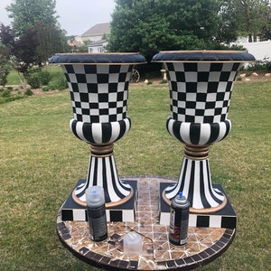 May include: Two black and white checkerboard patterned urns with gold accents on a black and white tiled table. The urns are on square bases with black and white checkerboard patterns. Two spray paint cans are on the table.