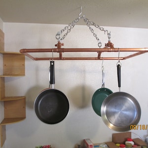 May include: A copper pipe pot rack suspended from the ceiling by chains, holding three frying pans with black and silver handles. A wooden corner shelf is visible on the left.