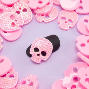 May include: A collection of pink skull-shaped embellishments, some with sparkling rhinestones, scattered on a light purple surface. One black skull-shaped embellishment with rhinestones is in the center. The skulls are a mix of solid and open designs.