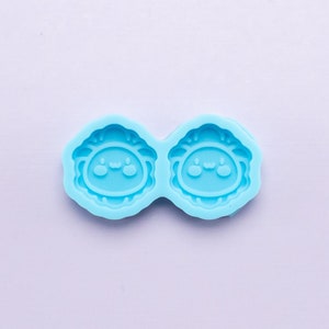 May include: A light blue silicone mold with two round cavities, each featuring a cartoon face with two eyes and a smiling mouth.