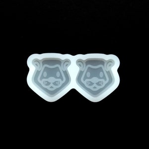 May include: A white silicone mold with two cavities shaped like animal faces. Each cavity features a detailed design of an animal with black eyes and a nose.