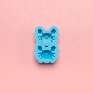 May include: Blue silicone mold with two crab shapes. The mold is on a pink background.