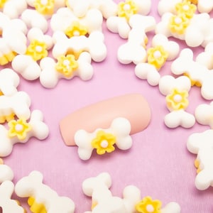 May include: White bone-shaped charms with yellow flower accents scattered on a pink surface. A single pink nail with a yellow flower accent is in the center of the image.