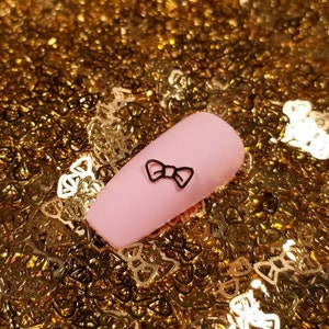 May include: A single pink artificial nail with a black bow design on a background of gold glitter.