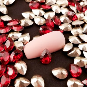 May include: A close-up of a pink nail with a variety of red and gold rhinestones scattered around it.