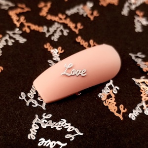 May include: Silver and rose gold metallic nail charms with the word "Love" written on them. The charms are scattered on a black surface with a fingernail painted pink and decorated with a silver "Love" charm.