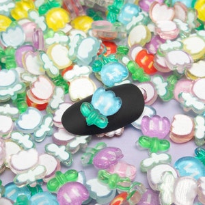 May include: A collection of colorful, translucent flower-shaped embellishments in various colors, including red, pink, yellow, and blue. The small, decorative pieces are scattered across a light purple surface, with a black oval object in the center.