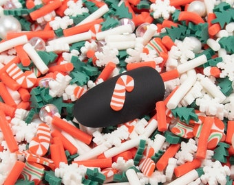 May include: A close-up of a variety of colorful sprinkles, including red and white candy canes, green trees, and white snowflakes. The sprinkles are scattered on a black surface.