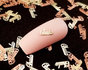 May include: A pink nail with the word "Chula" written on it. The nail is surrounded by gold and rose gold confetti with the same word written on each piece.