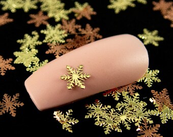 May include: A close-up of a fingernail with a gold snowflake nail art design. The nail is surrounded by gold and copper snowflake confetti on a black background.