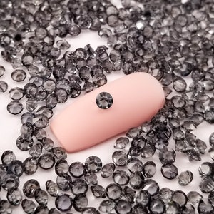 May include: A collection of small, faceted, gray rhinestones scattered across a white surface. One rhinestone is placed on a light pink nail, showcasing the size and color of the gems. The rhinestones are round and appear to be made of glass or crystal.