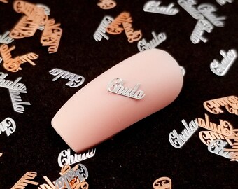 May include: A close-up of a pink nail with a silver metal accent that says "Chula" on a black background. The background is covered in silver and rose gold metal accents that also say "Chula".