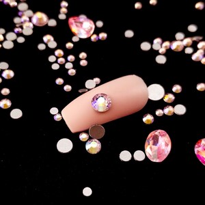 May include: A close-up of a light pink nail with a clear iridescent rhinestone on top. The nail is surrounded by a scattering of small, clear iridescent rhinestones on a black background.
