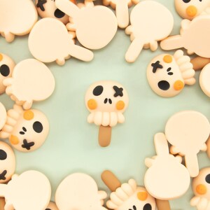 May include: A collection of small, beige, skull-shaped charms with black eyes and mouths. The charms have a textured surface and are scattered on a light green background.