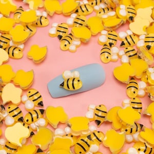 May include: A close-up of a light blue nail file with a yellow and black bee charm on top. The nail file is surrounded by a variety of yellow and black bee-shaped charms on a pink background.