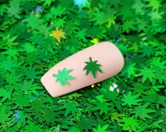 May include: A close-up of a fingernail with two green marijuana leaf nail art stickers on a bed of green glitter.