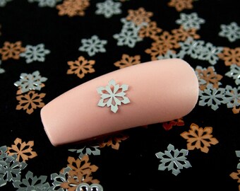 May include: A pink false nail with a silver snowflake design on a black background surrounded by silver and copper snowflake confetti.
