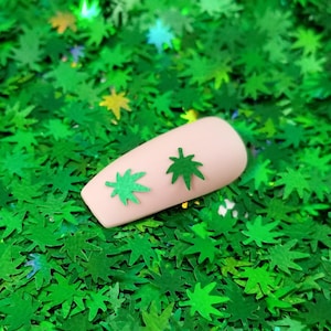 May include: A close-up of a fingernail with two green marijuana leaf nail art stickers on a bed of green glitter.