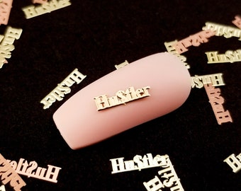May include: A close-up of a pink nail with a gold metal accent that says "Hustler". The nail is surrounded by many other gold metal accents that say "Hustler" in various sizes and orientations.