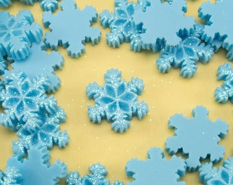 May include: A collection of light blue snowflake-shaped embellishments with glitter, scattered on a yellow surface.