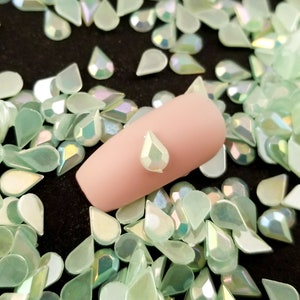 May include: A close-up of a light green iridescent teardrop-shaped rhinestone on a fingernail. The rhinestone is surrounded by a scattering of similar rhinestones.
