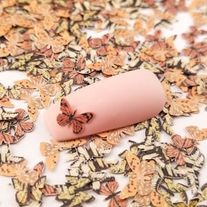 May include: A close-up of a pink nail with a small orange butterfly sticker on it. The nail is surrounded by a scattering of small, printed butterfly stickers in various shades of orange, yellow, and brown.