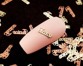May include: A collection of metallic nail art decorations spelling the name "Tricia" in a cursive font. The nail art is scattered on a black surface, with one piece placed on a pink nail. The decorations are in gold and rose gold tones.