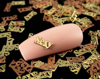 May include: A pink nail with a gold metal nail art accent that says "Sexy". The nail is surrounded by gold and rose gold metal confetti that also says "Sexy".