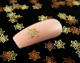 May include: A close-up of a pink nail with a gold snowflake design. The nail is surrounded by a scattering of gold and copper snowflake embellishments on a black background.