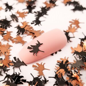 May include: Black and copper witch silhouette nail art embellishments. The embellishments are scattered on a white surface with a fingernail painted pink with a witch silhouette on it.
