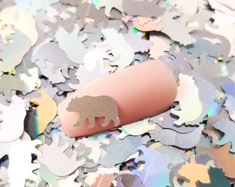 May include: A close-up of iridescent holographic glitter in the shape of bears. The glitter is scattered on a surface and a fingernail with a light pink polish is shown in the center of the image.