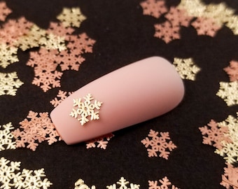 May include: A close-up of a pink nail with a gold snowflake nail art design. The nail is surrounded by a scattering of gold and rose gold snowflake confetti.