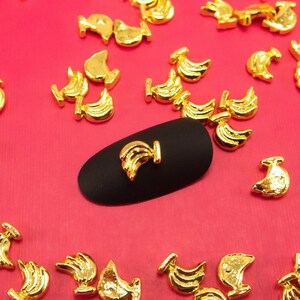 May include: Gold-coloured metal nail art charms in the shape of a stylised bird. The charms are scattered on a pink surface with a black nail art tool in the centre.